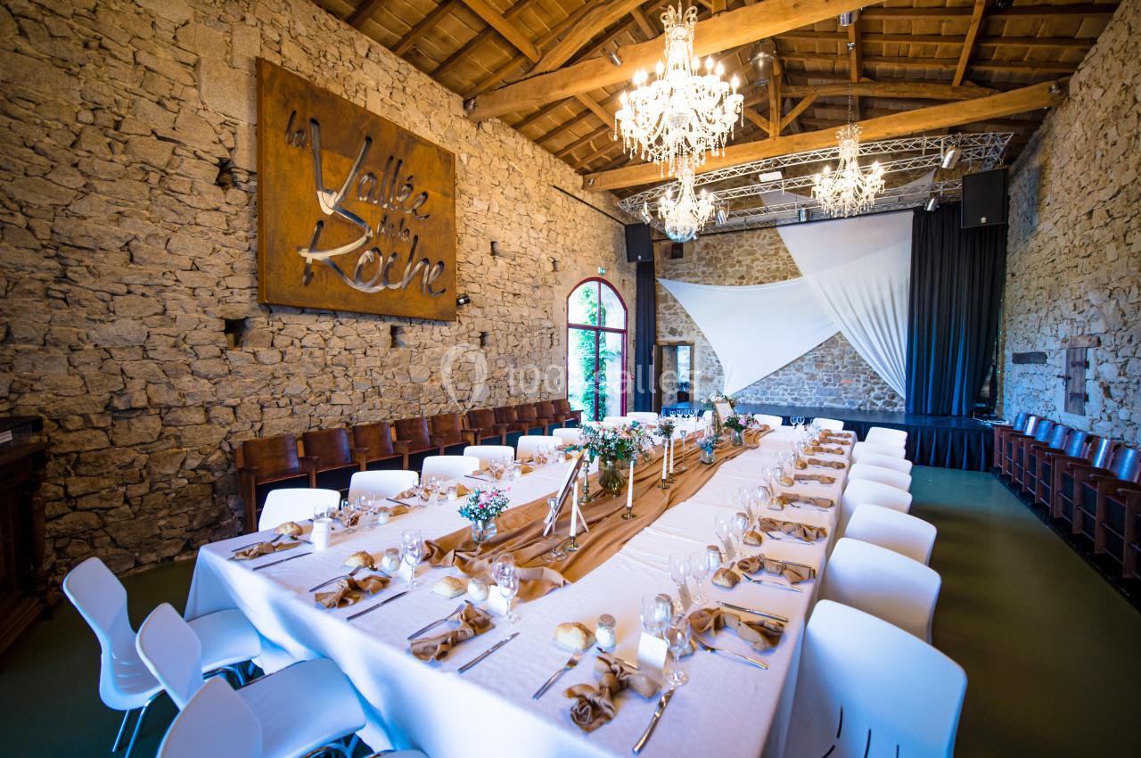 Salle en pierre avec une grande table dressée pour un repas, décorée de fleurs et éclairée par des lustres.