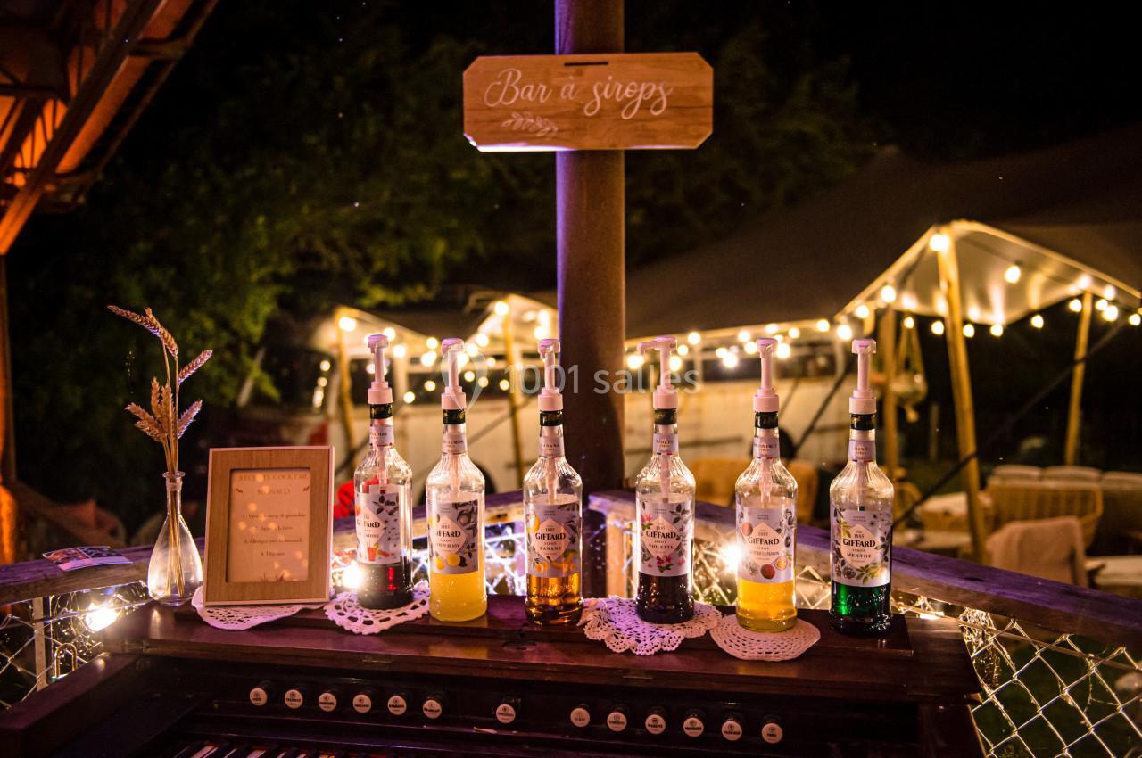 Bouteilles de sirops alignées sur un comptoir en bois, éclairées par des guirlandes lumineuses dans un cadre nocturne.