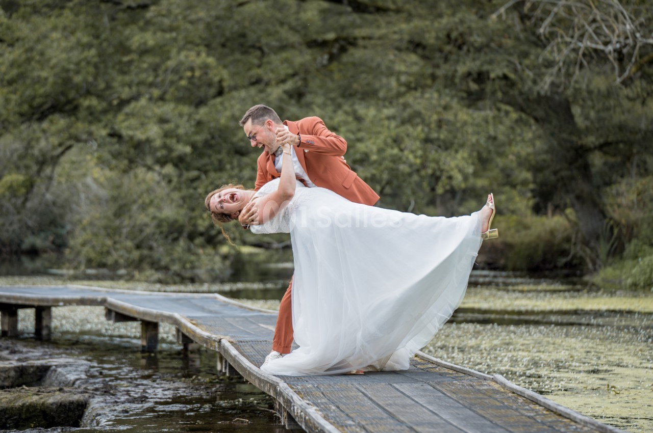 Un couple en tenue de mariage danse sur un pont en bois au-dessus d'un plan d'eau entouré de végétation.