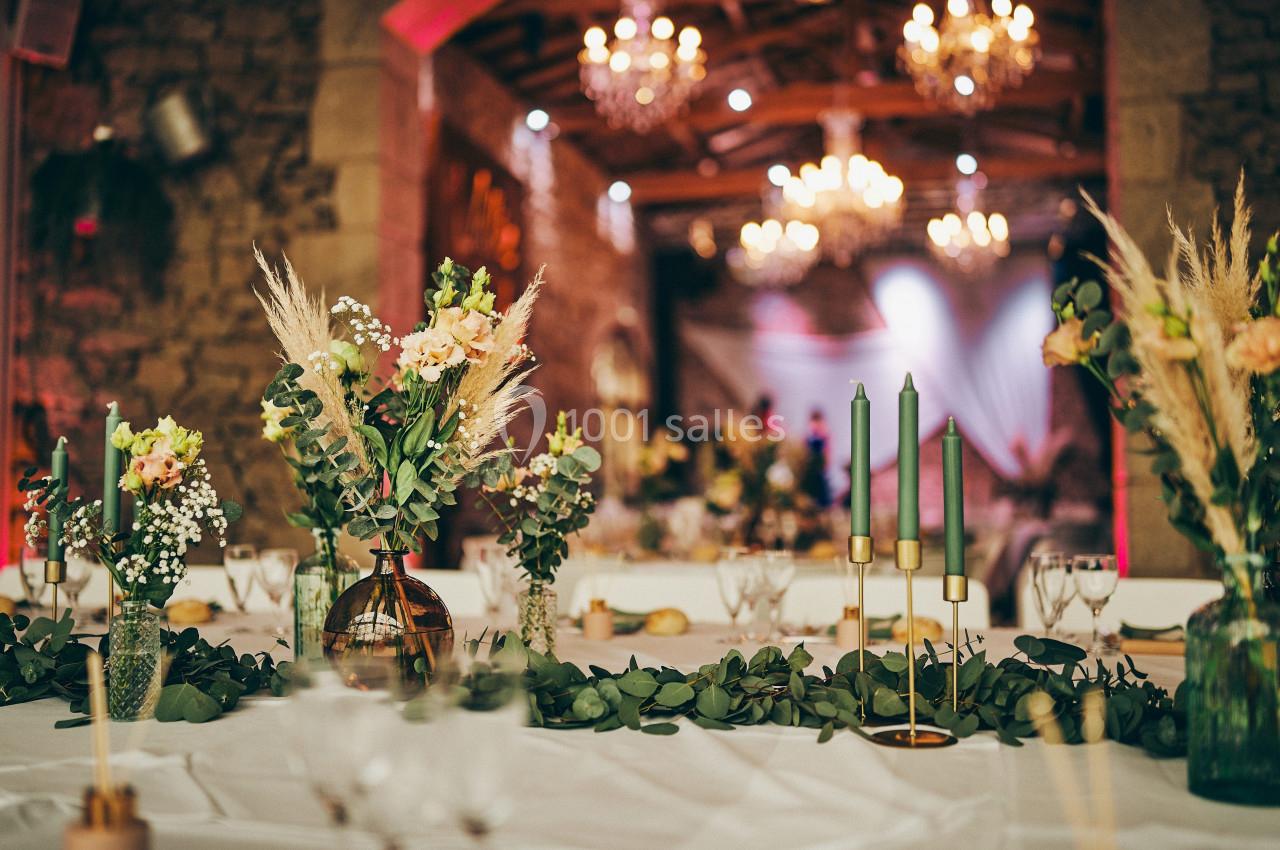 Table décorée avec des bouquets de fleurs, chandeliers verts et guirlande végétale dans une salle éclairée par des lustres.