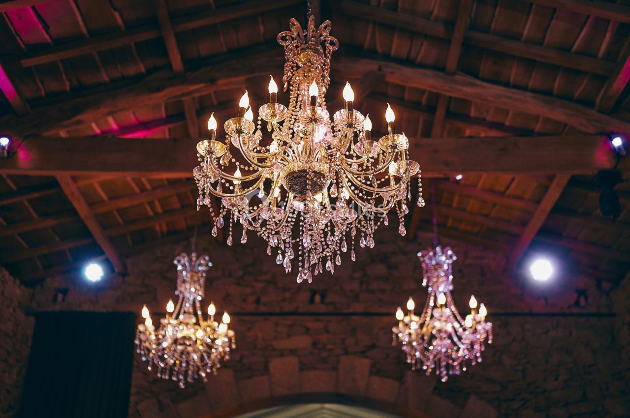 Trois lustres en cristal éclairés suspendus dans une salle aux murs en pierre et au plafond en bois.