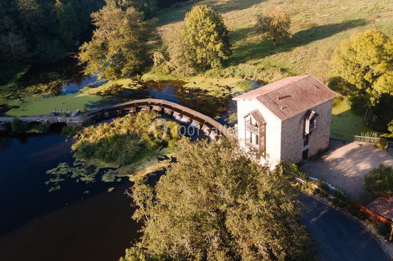 Vue aérienne d'un moulin en pierre près d'une rivière avec un pont en bois entouré de végétation.