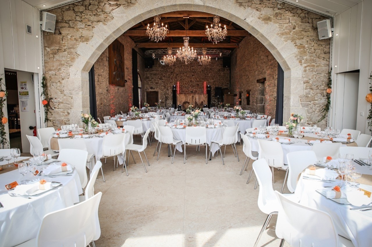 Salle de réception en pierre avec tables rondes dressées pour un événement, décorations florales et lustres suspendus.