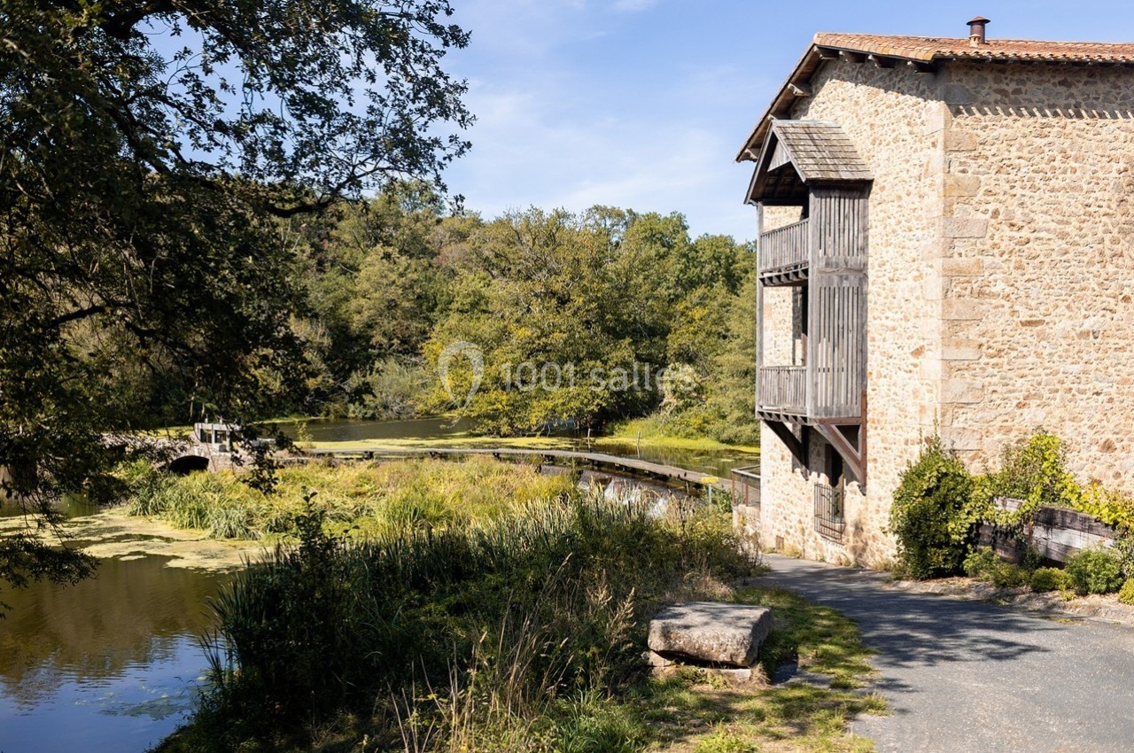 Bâtiment en pierre au bord d'une rivière entourée de végétation, avec un pont en arrière-plan sous un ciel dégagé.