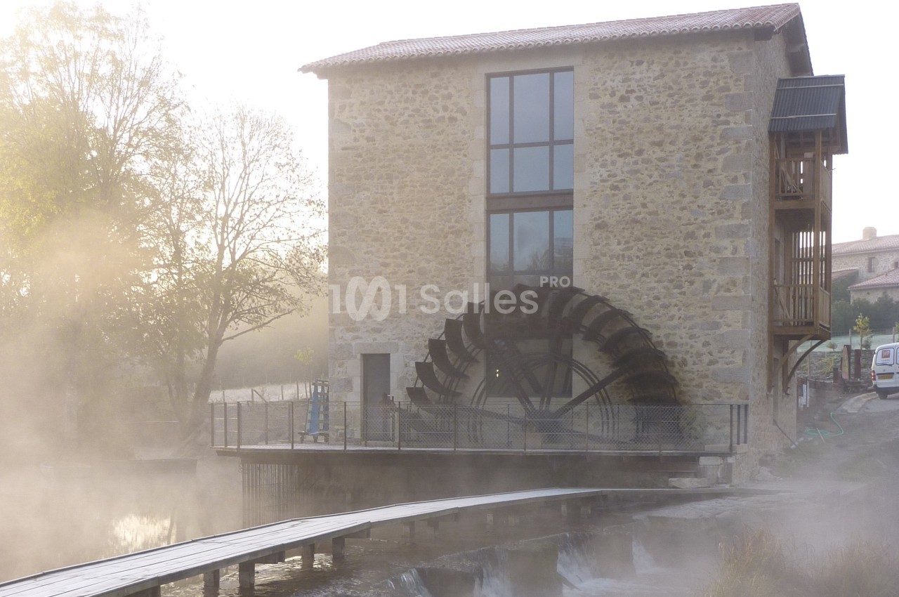 Bâtiment en pierre avec une grande roue à aubes, entouré de brume et bordé d'un cours d'eau.