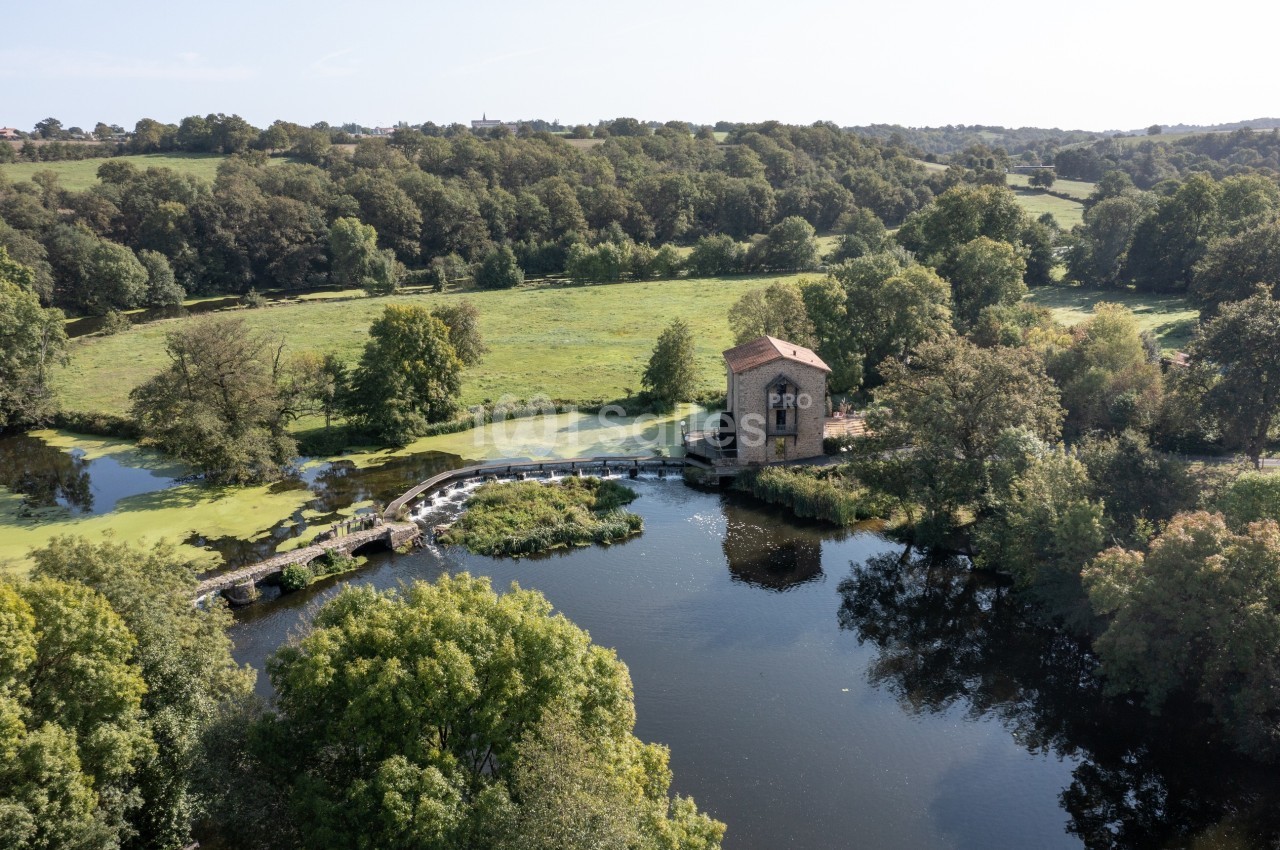 Vue aérienne d'un moulin en pierre près d'une rivière entourée de verdure et de collines boisées.