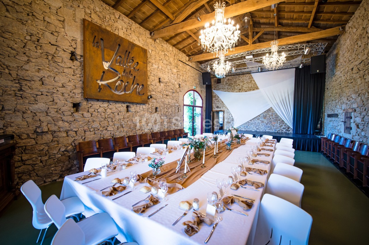 Salle de réception en pierre avec une grande table dressée, décorée de fleurs et éclairée par des lustres.