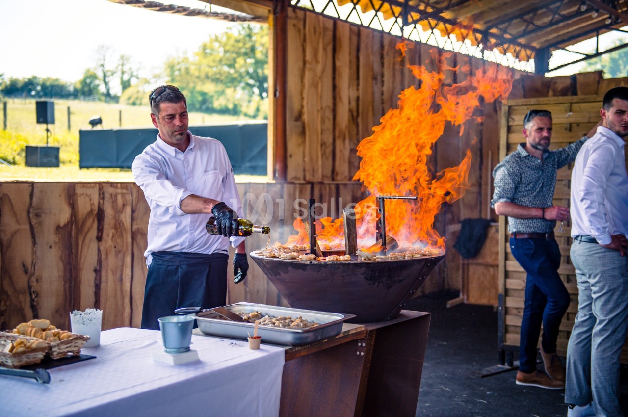 Un chef verse du vin sur une grande poêle enflammée lors d'une cuisson en extérieur sous un abri en bois.