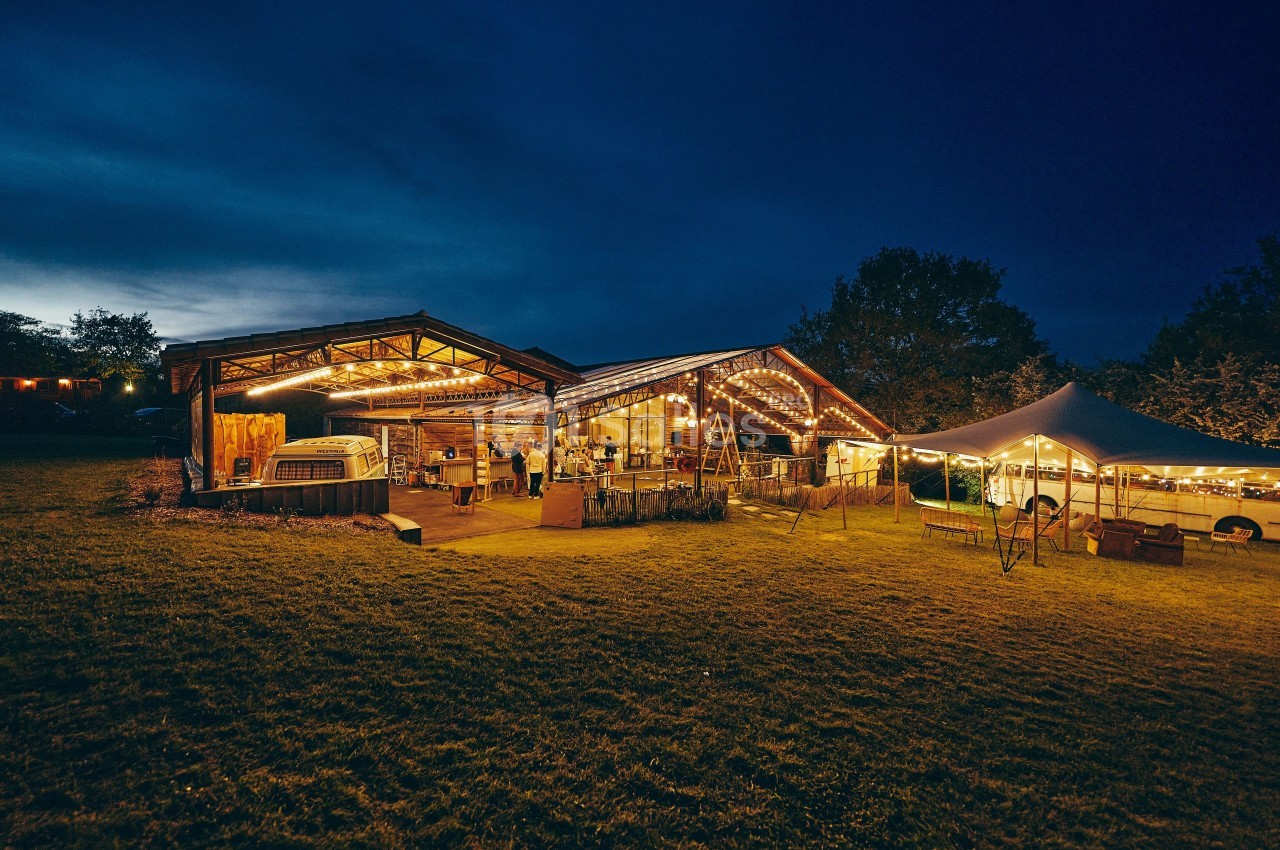 Grange éclairée en soirée avec des guirlandes lumineuses, entourée d'une pelouse et d'une tente blanche.