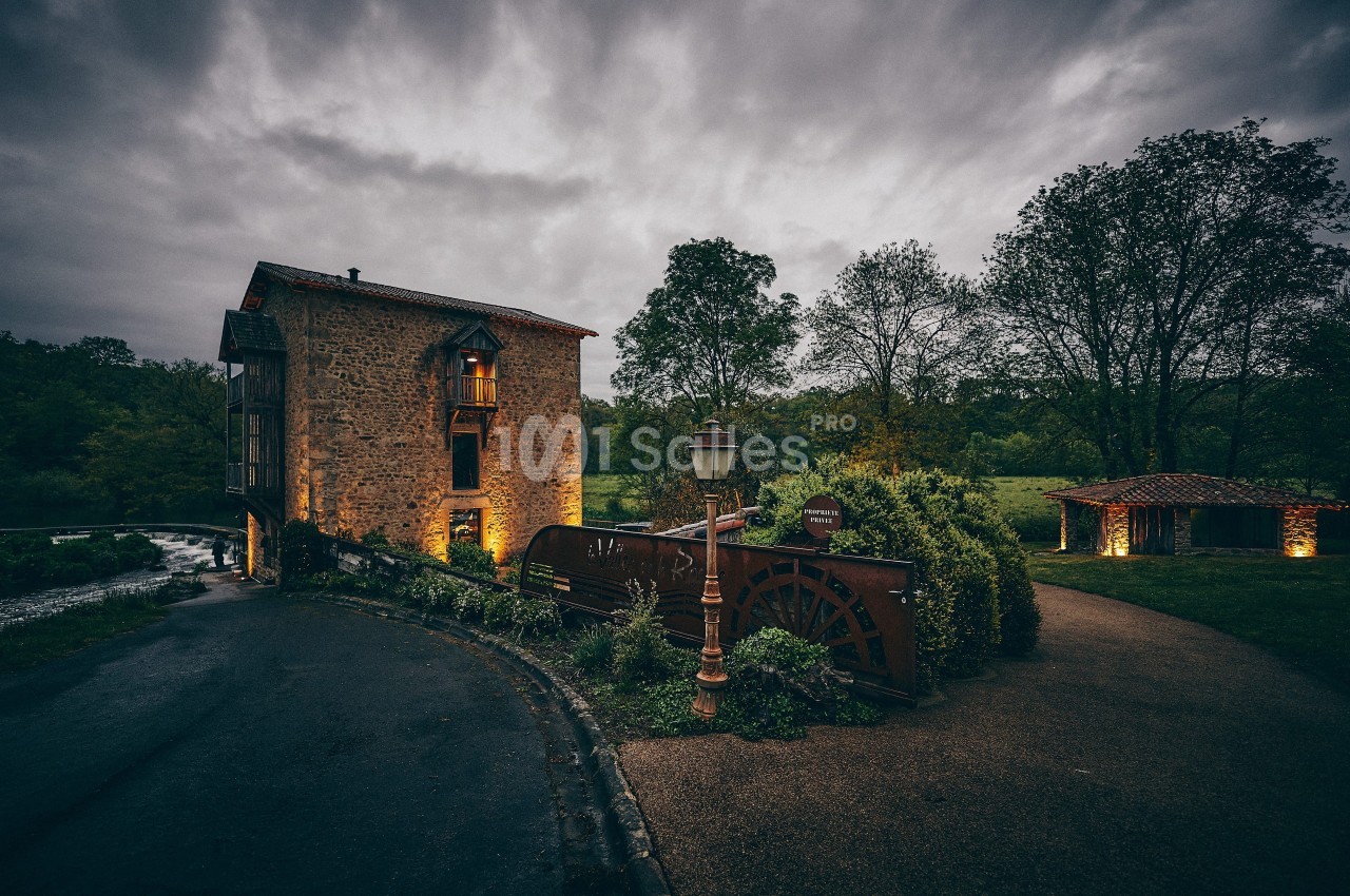 Ancien moulin en pierre avec roue à aubes, éclairé au crépuscule, entouré de verdure et d'un chemin pavé.