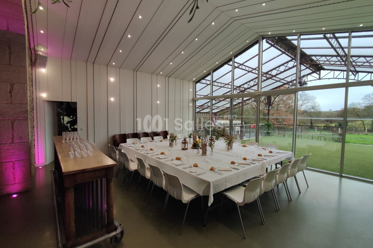 Salle lumineuse avec grandes baies vitrées, tables dressées pour un repas, décor sobre et vue sur un espace extérieur…