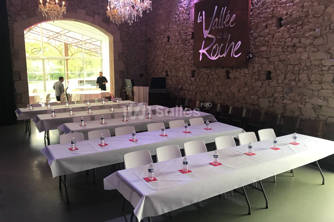 Salle de réception avec tables alignées, nappes blanches, bouteilles d'eau et lumière naturelle entrant par une baie vitrée.