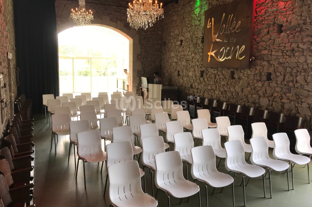 Salle en pierre avec rangées de chaises blanches, lustres suspendus et une grande baie vitrée en arrière-plan.