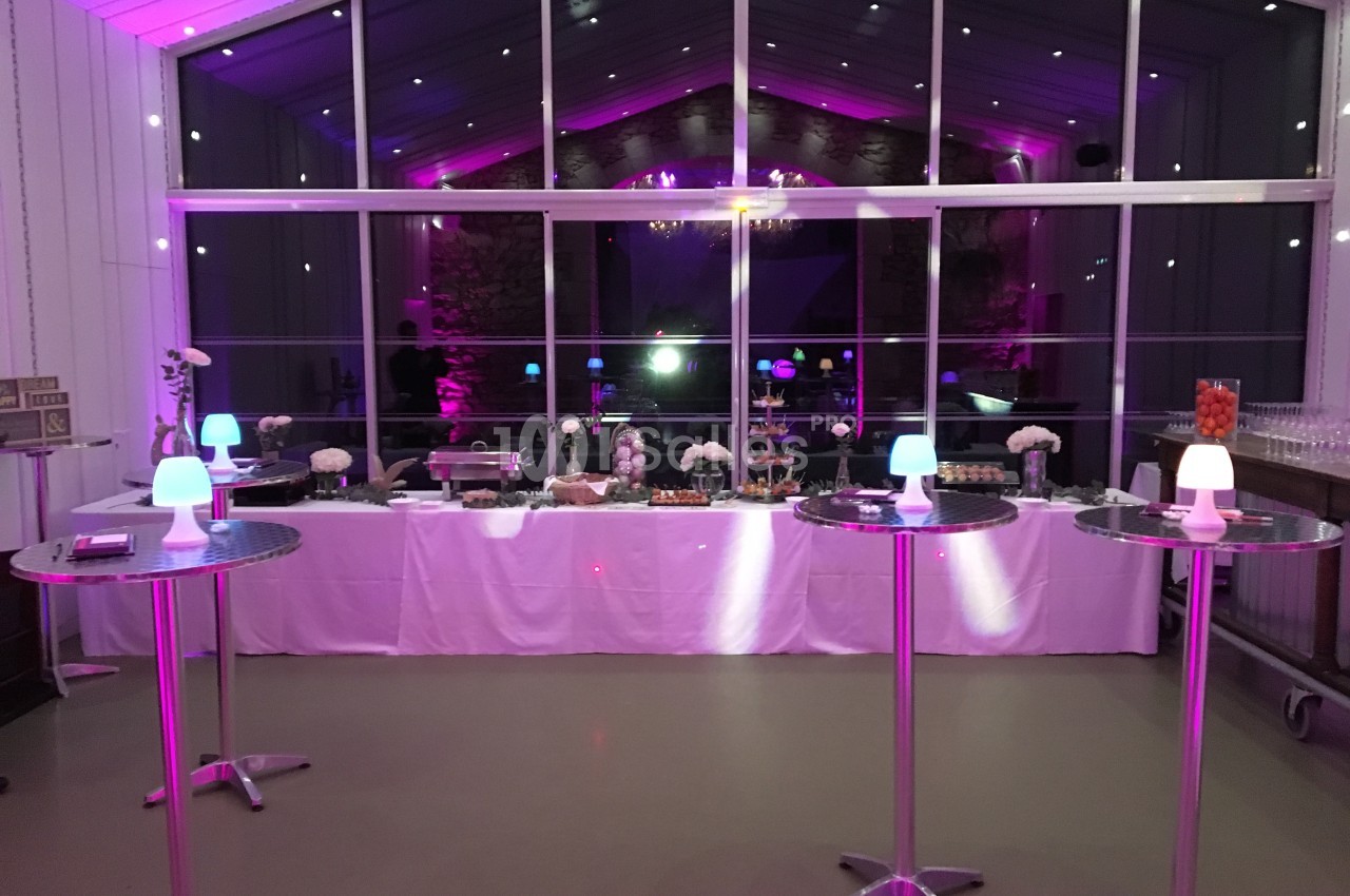 Salle de réception éclairée en rose avec un buffet dressé et des tables hautes décorées de petites lampes.