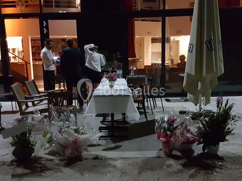 Table dressée en extérieur avec des bouquets de fleurs au sol, devant un bâtiment éclairé en soirée.