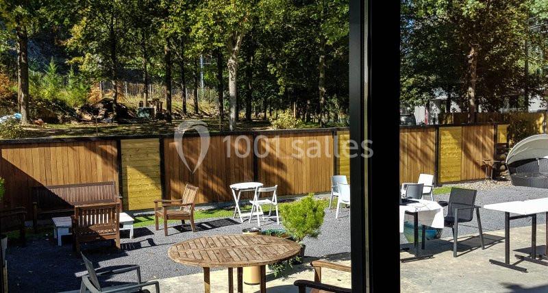 Vue d'une terrasse avec des tables, des chaises et une clôture en bois, entourée d'arbres sous un ciel ensoleillé.