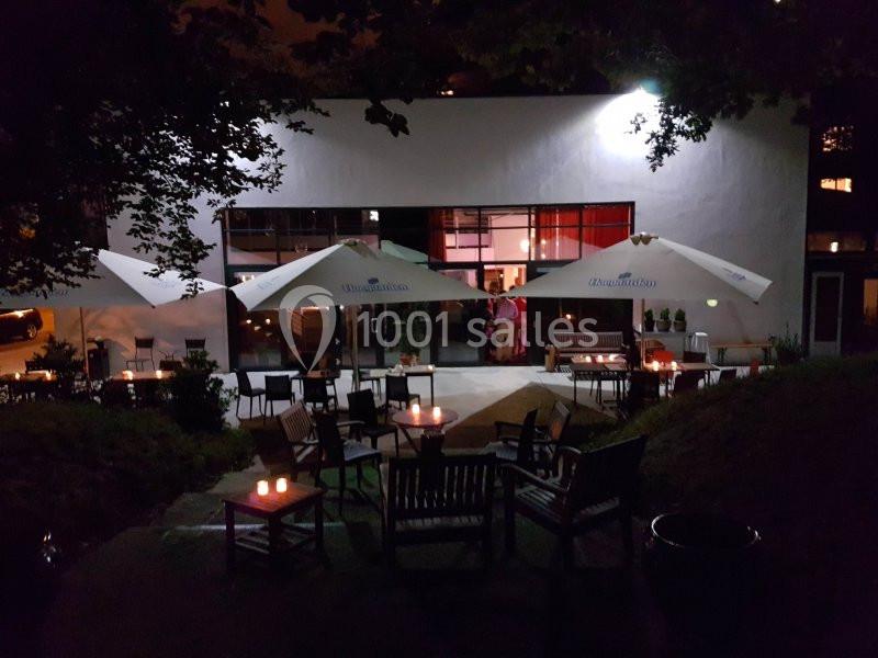 Terrasse éclairée d'un restaurant avec tables, chaises et parasols, vue de nuit sous des arbres.
