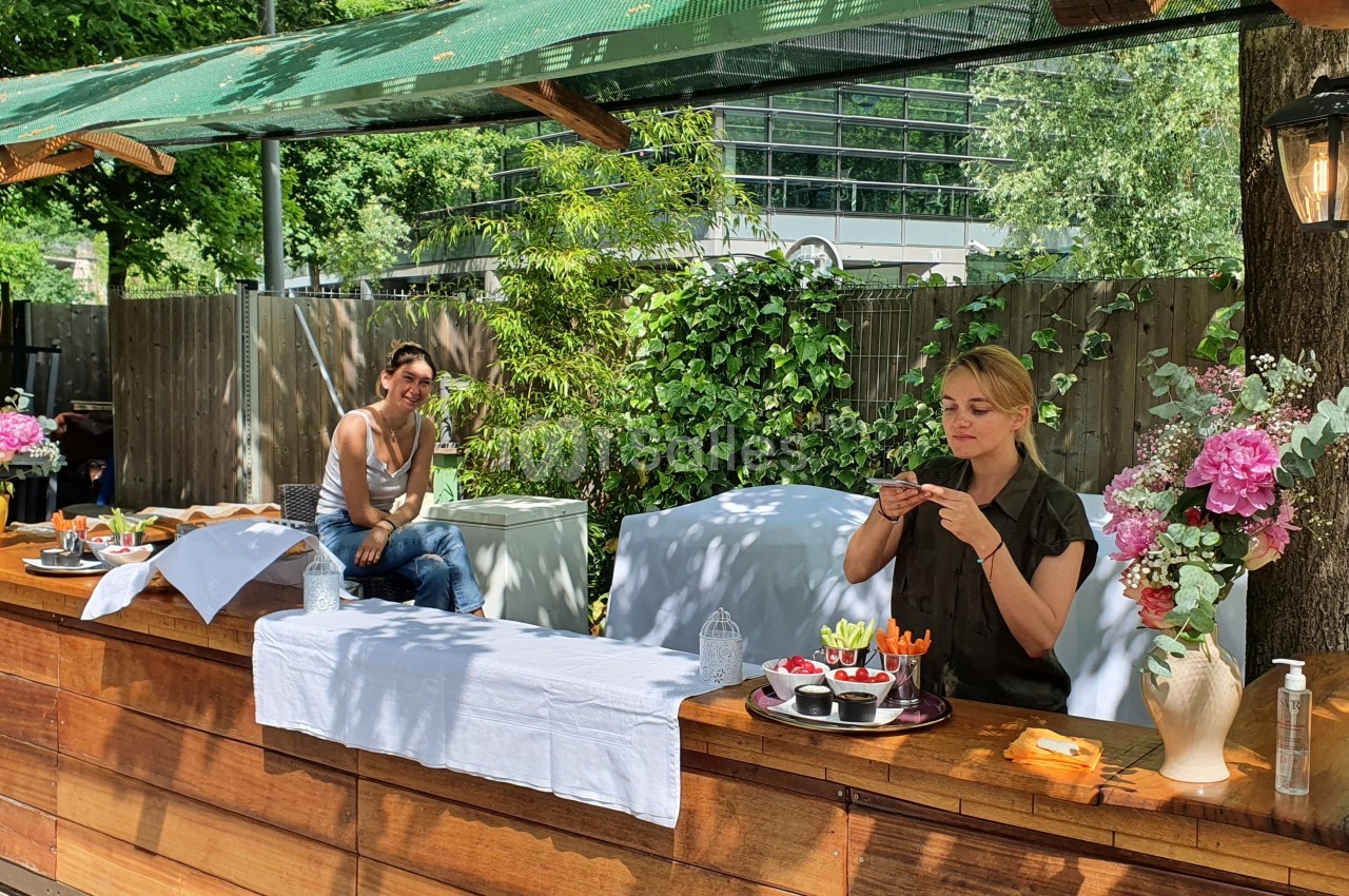 Deux femmes dans un stand en bois en extérieur, l'une préparant des plats, l'autre assise en arrière-plan.