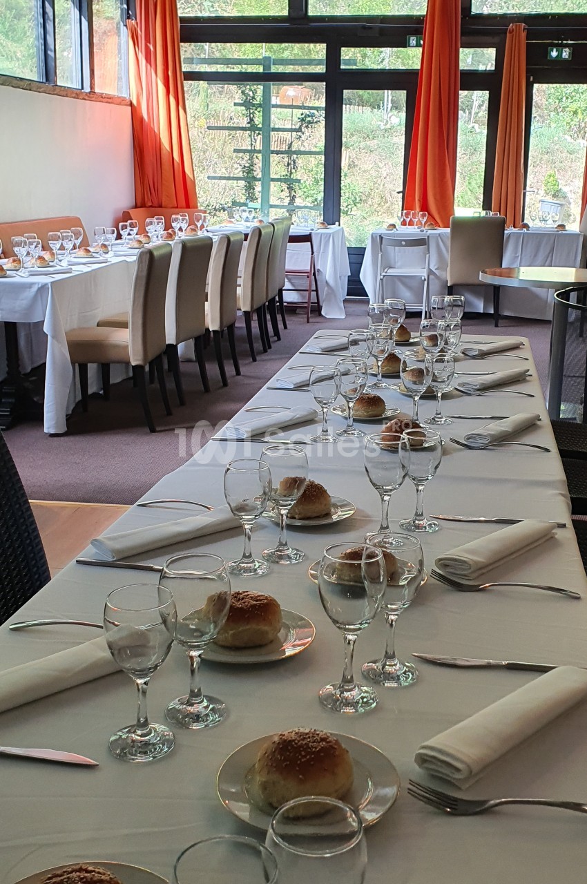 Salle de réception lumineuse avec tables dressées, nappes blanches, verres, assiettes et petits pains alignés.