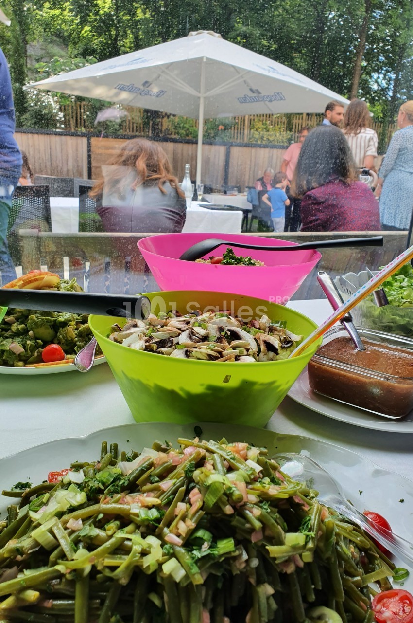 Buffet avec salades variées en premier plan, terrasse extérieure avec convives en arrière-plan sous des parasols.