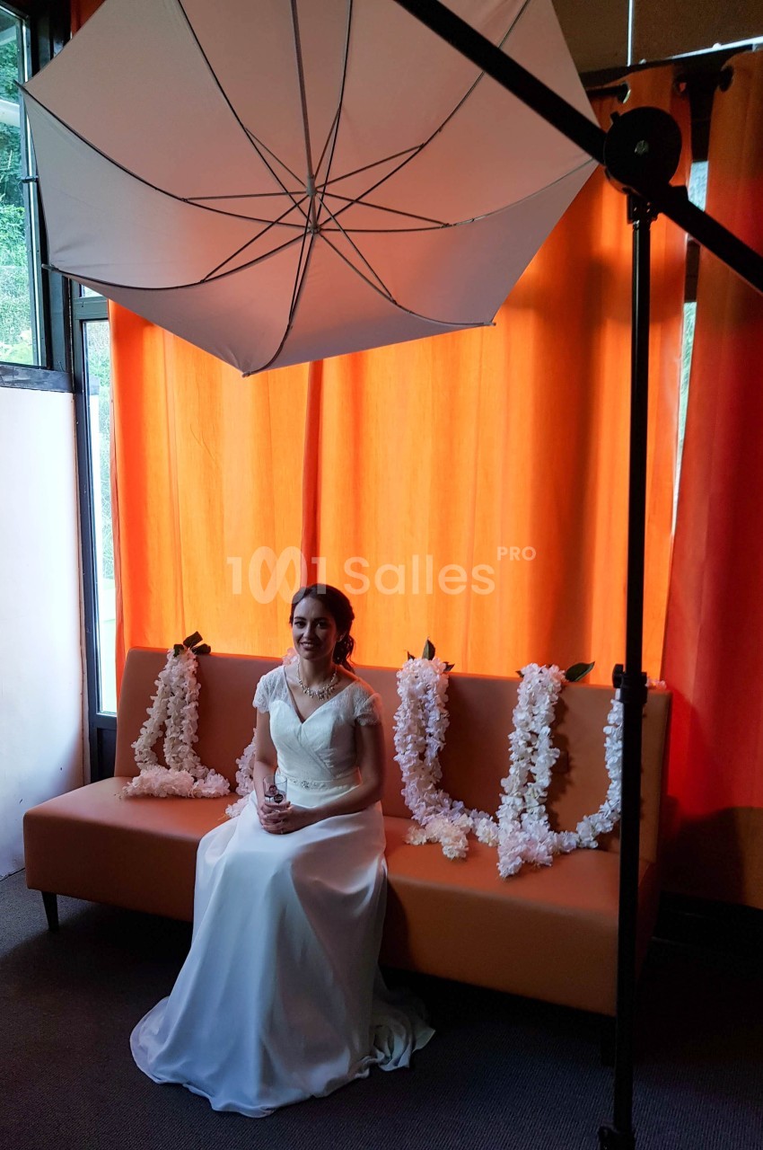 Une femme en robe de mariée est assise sur un canapé, éclairée par un parapluie de studio devant des rideaux orange.