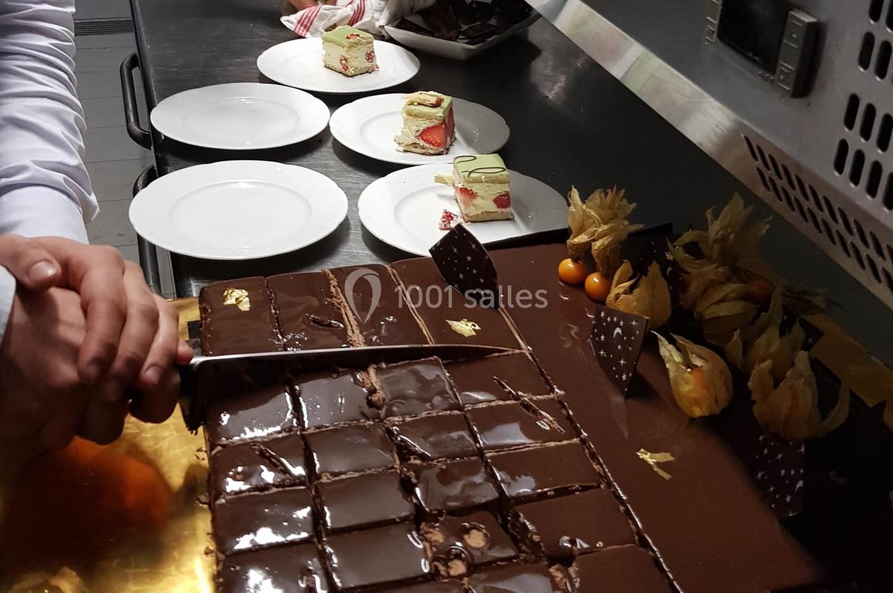 Découpe d'un gâteau au chocolat brillant en cuisine, avec des desserts et assiettes préparés en arrière-plan.