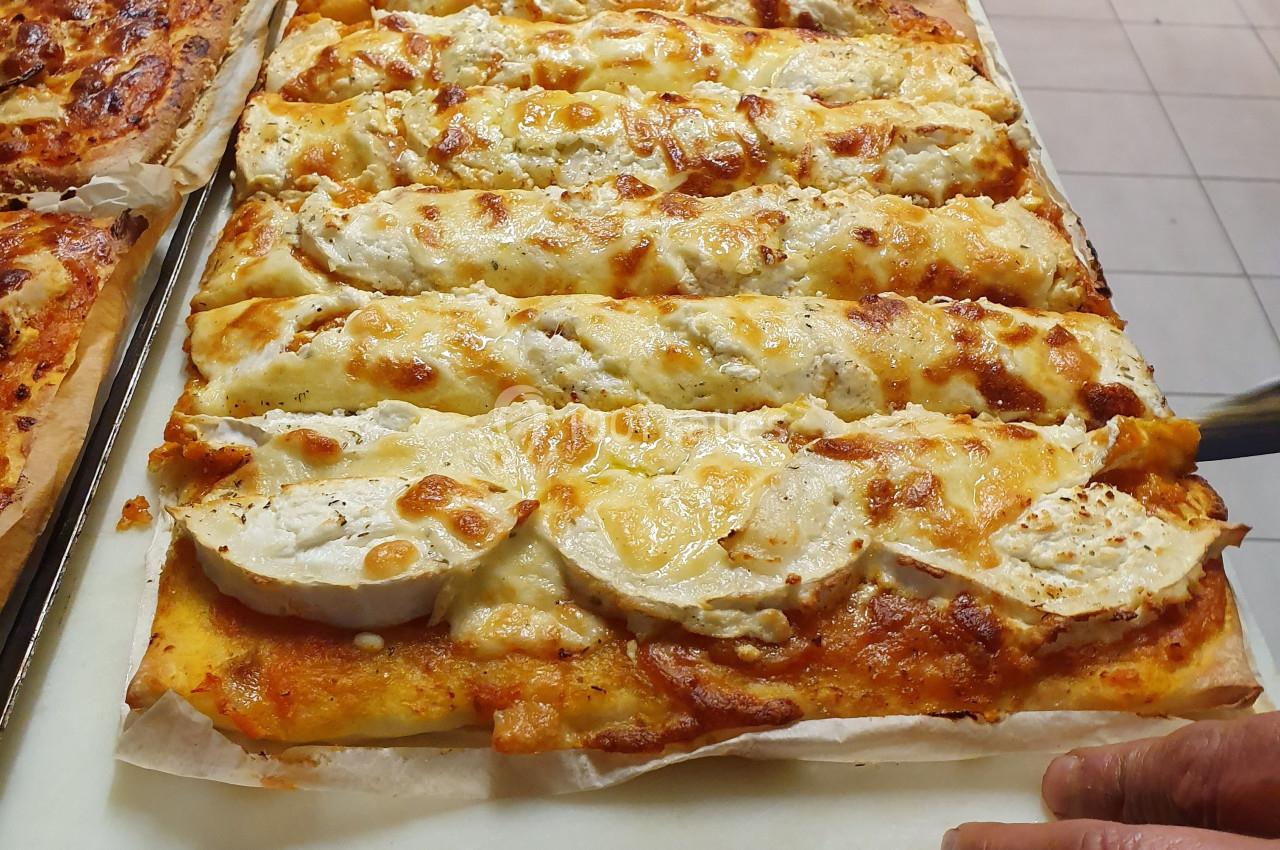 Pizza rectangulaire garnie de fromage de chèvre et de sauce tomate, posée sur une planche dans une cuisine.