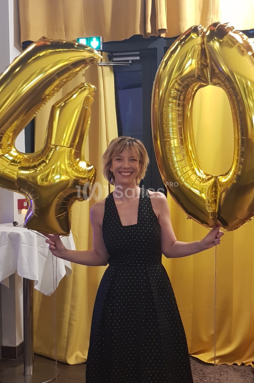 Femme souriante tenant des ballons dorés formant le chiffre 40 dans une salle décorée de rideaux jaunes.
