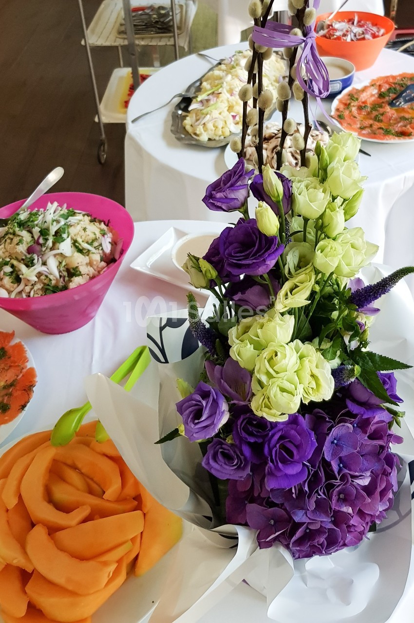 Buffet avec des plats variés, dont des salades, du saumon fumé et des fruits, décoré d'un bouquet de fleurs violettes et…