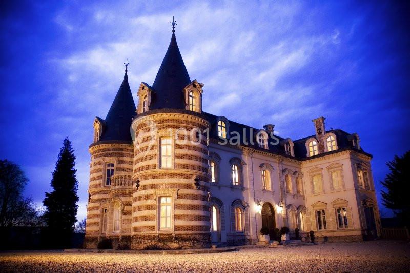Façade d'un château éclairé au crépuscule, avec des tours et un ciel bleu nuageux en arrière-plan.