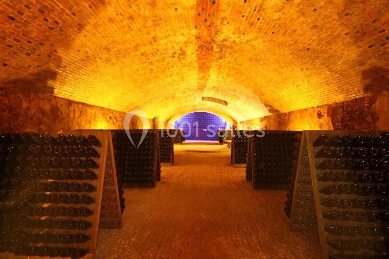 Cave voûtée en briques avec des bouteilles de vin stockées sur des supports en bois, éclairage chaud.