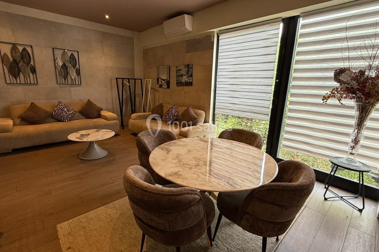 Salon moderne avec canapés, table basse, table ronde en marbre, chaises rembourrées et grande fenêtre avec stores.
