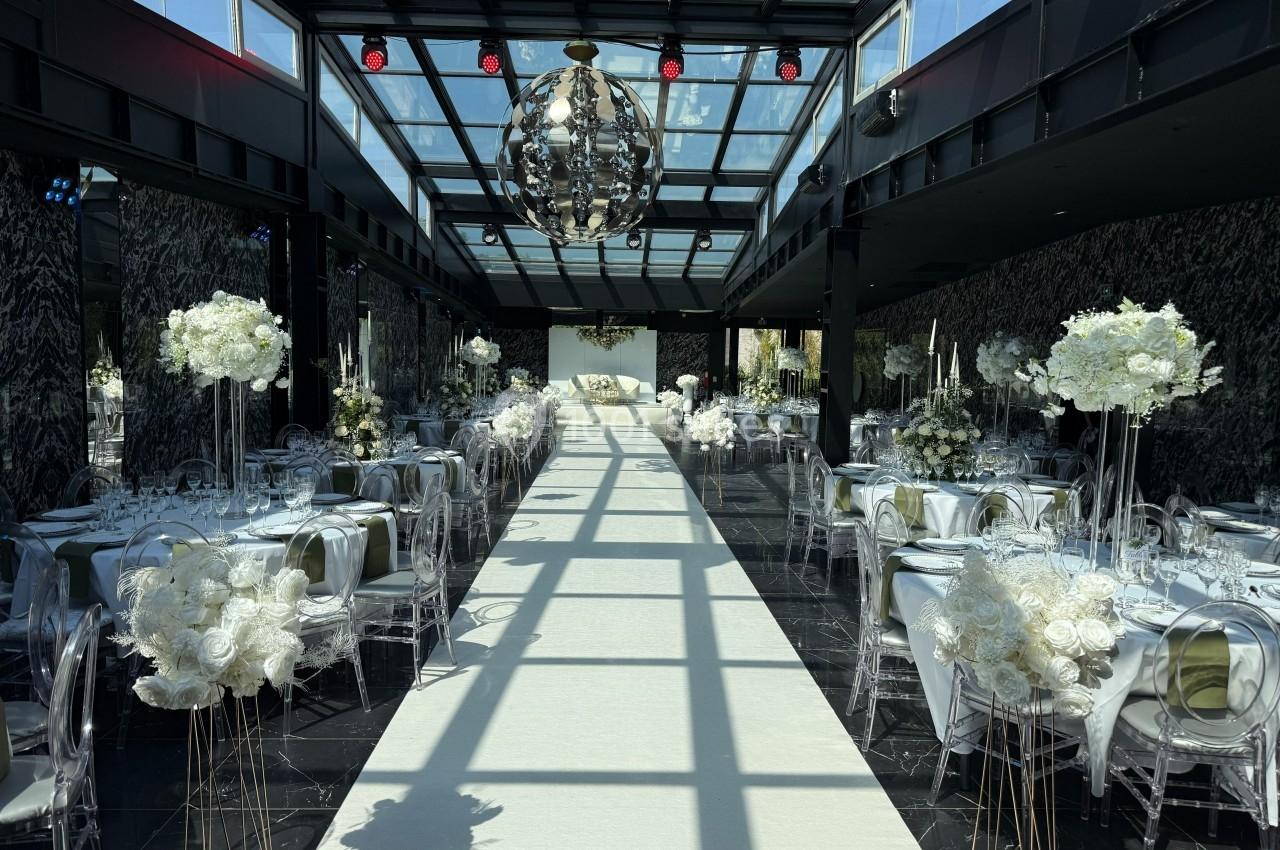 Salle de réception élégante avec tables décorées de fleurs blanches et un long tapis blanc sous une verrière.