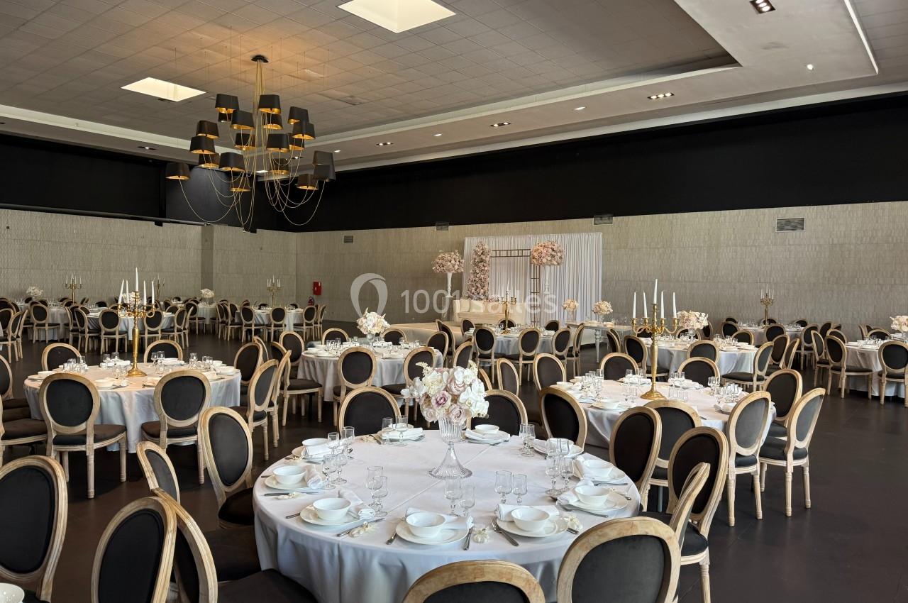 Salle de réception élégante avec tables rondes dressées, chaises en bois, décor floral et éclairage suspendu moderne.