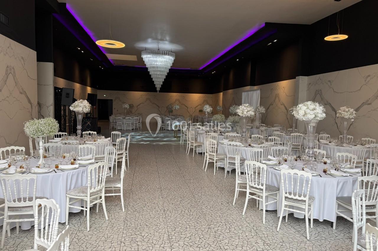 Salle de réception élégante avec tables rondes dressées, chaises blanches et décorations florales blanches.