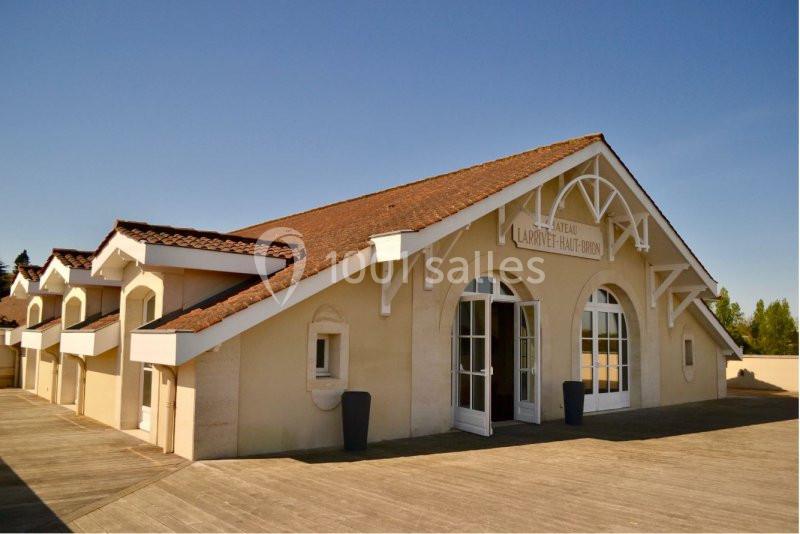Bâtiment clair avec toit en tuiles rouges, grandes arches vitrées et terrasse en bois sous un ciel dégagé.