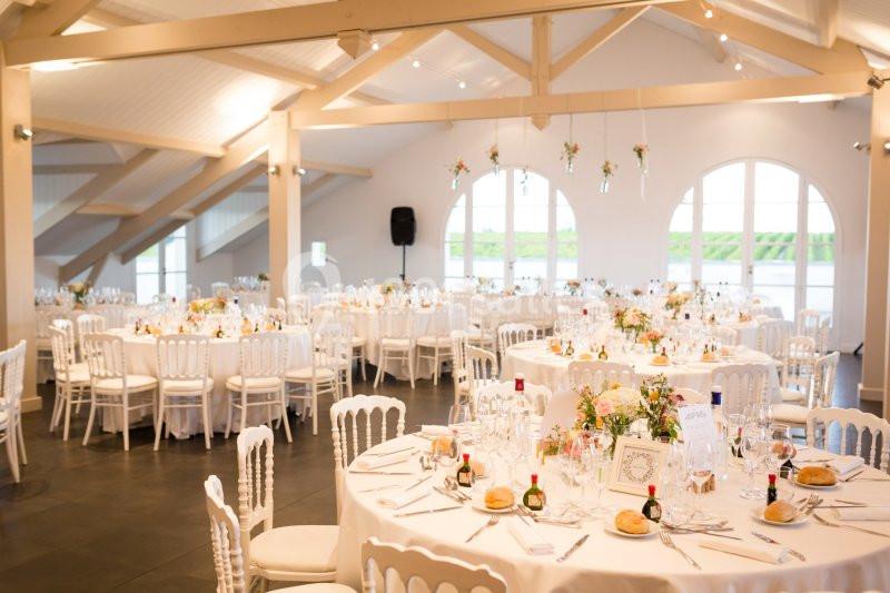 Salle lumineuse avec tables rondes décorées pour un événement, chaises blanches et grandes fenêtres en arrière-plan.