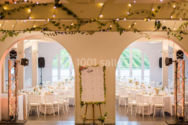 Salle de réception décorée avec des guirlandes lumineuses, des tables dressées et un plan de table sur chevalet.