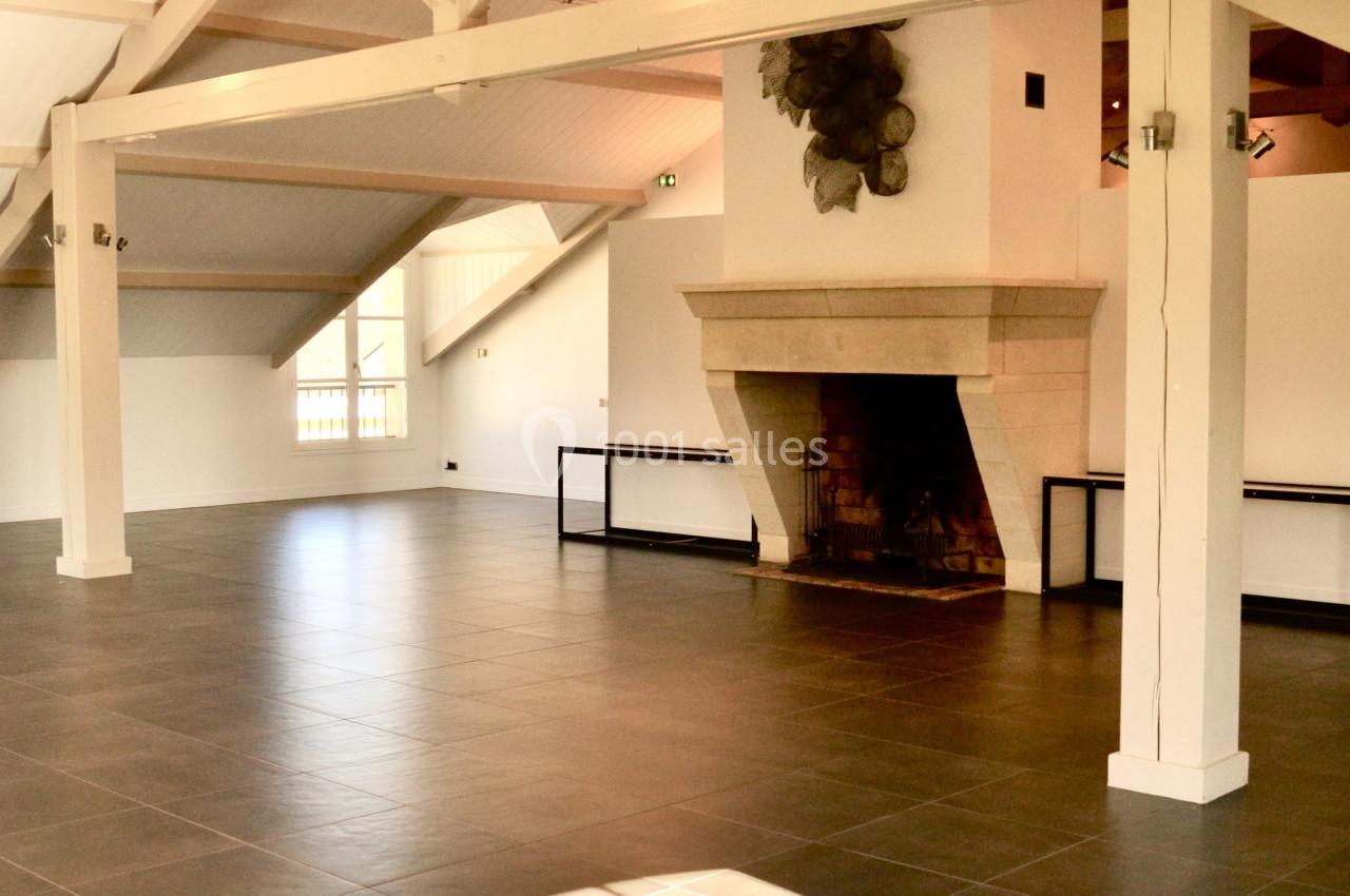 Grande pièce lumineuse avec poutres apparentes, cheminée en pierre et sol en carrelage sombre.