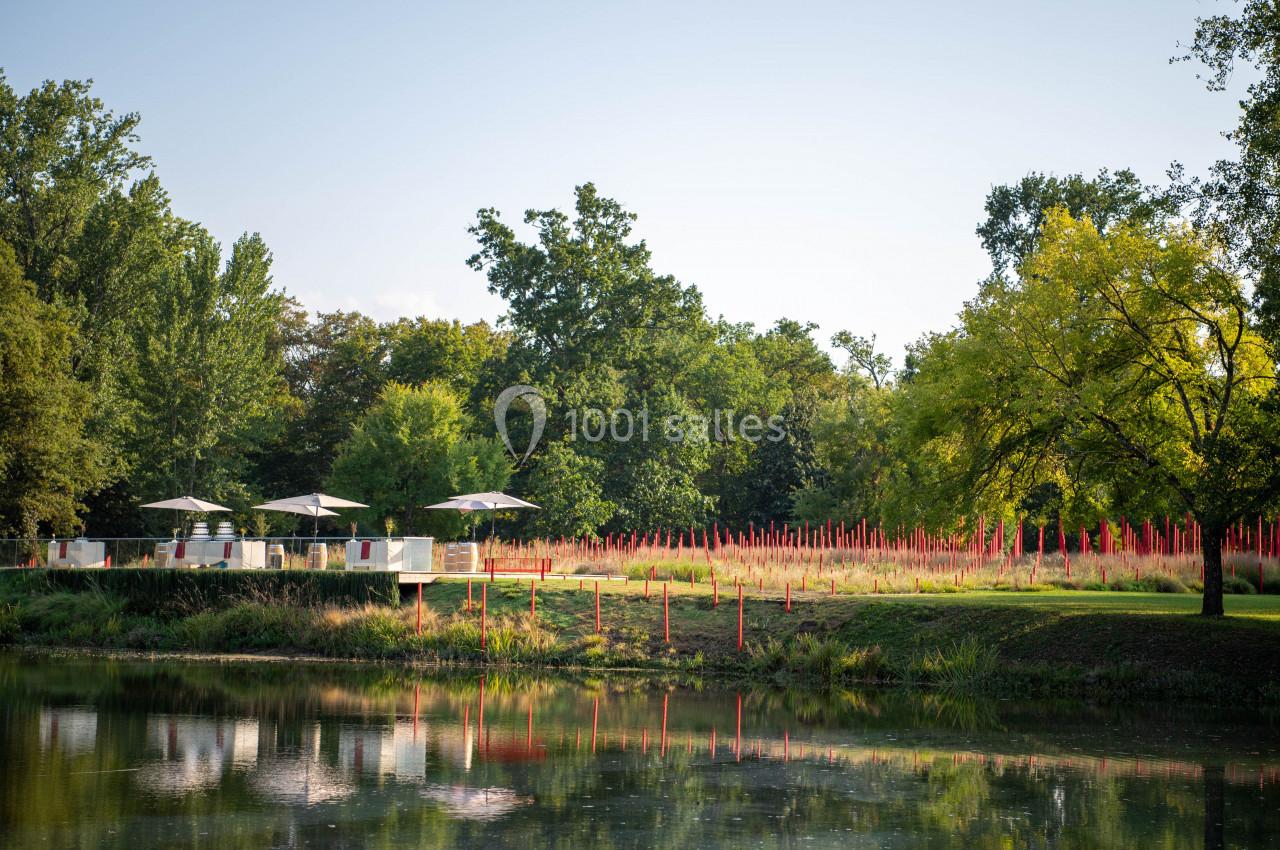Terrasse avec parasols blancs près d'un étang, entourée de végétation et de hautes herbes rouges.