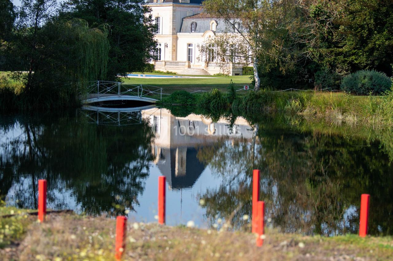 Manoir blanc entouré d'arbres, reflété dans un étang, avec des piquets rouges au premier plan.