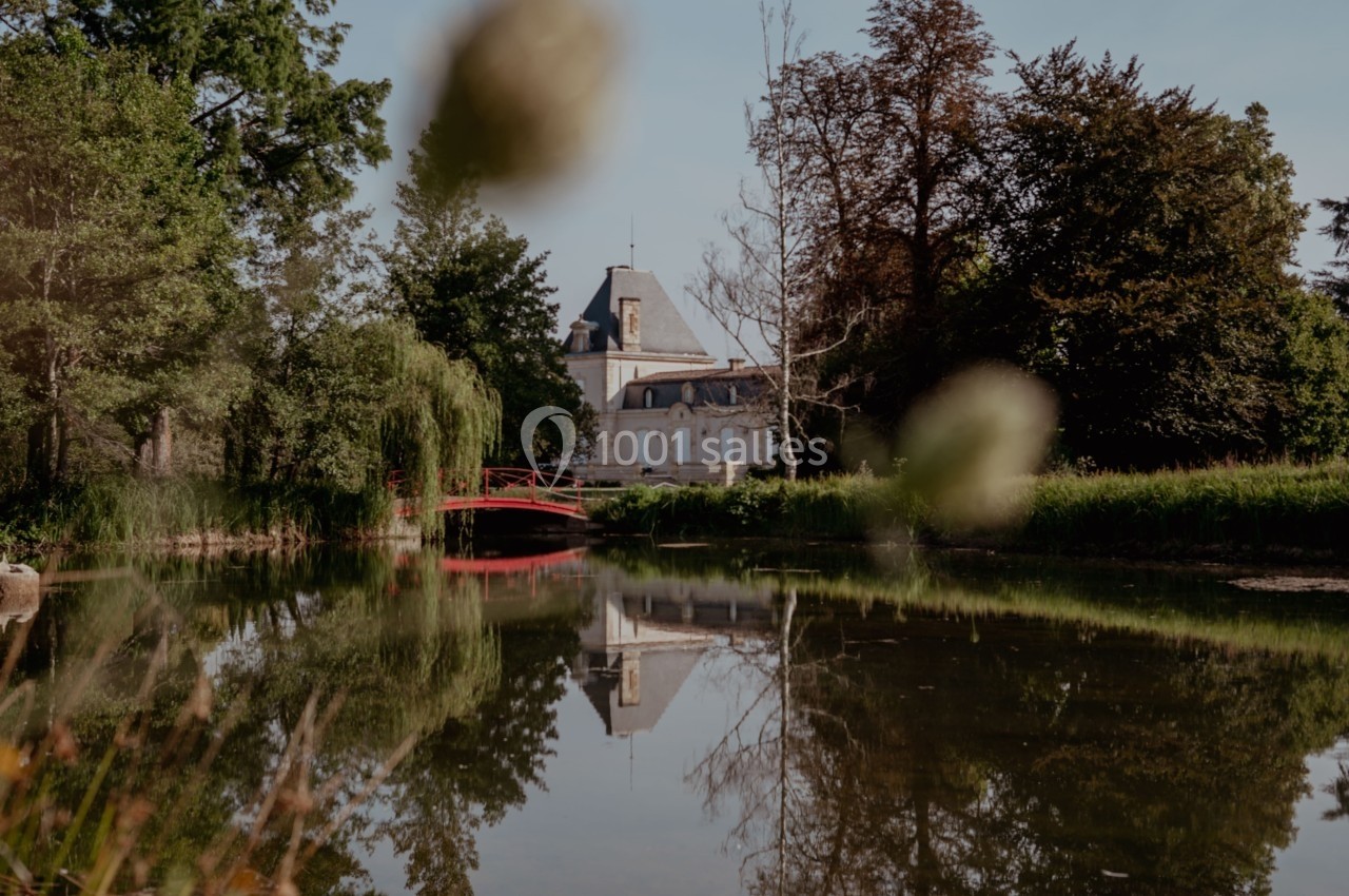 Vue d'un château au bord d'un étang entouré d'arbres, avec un pont rouge reflété dans l'eau calme.