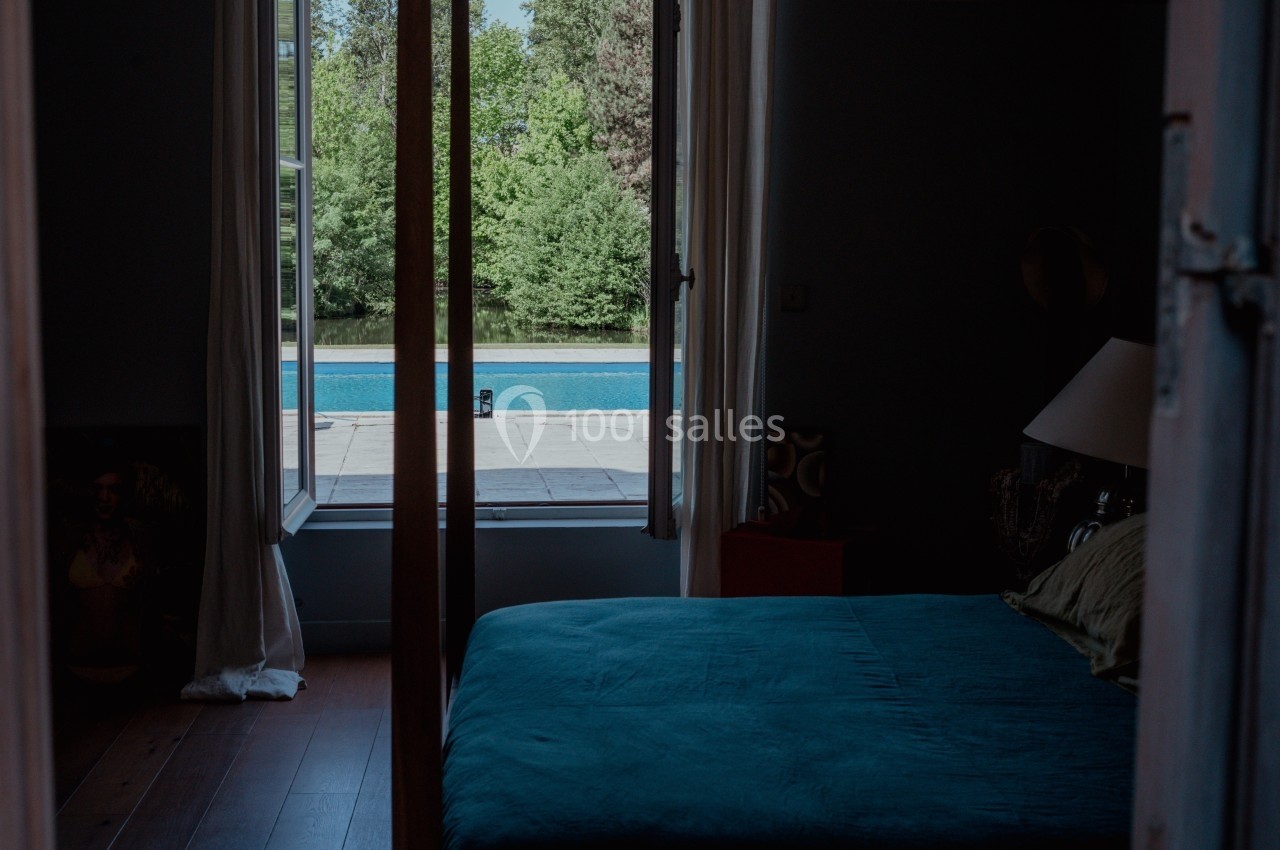 Vue depuis une chambre sombre avec lit bleu, donnant sur une terrasse et une piscine entourée de verdure.