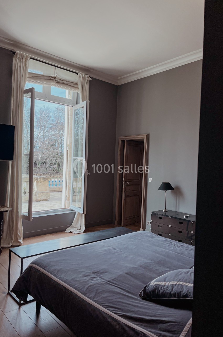 Chambre avec lit double, grande fenêtre ouverte sur un jardin, murs sombres et mobilier minimaliste.