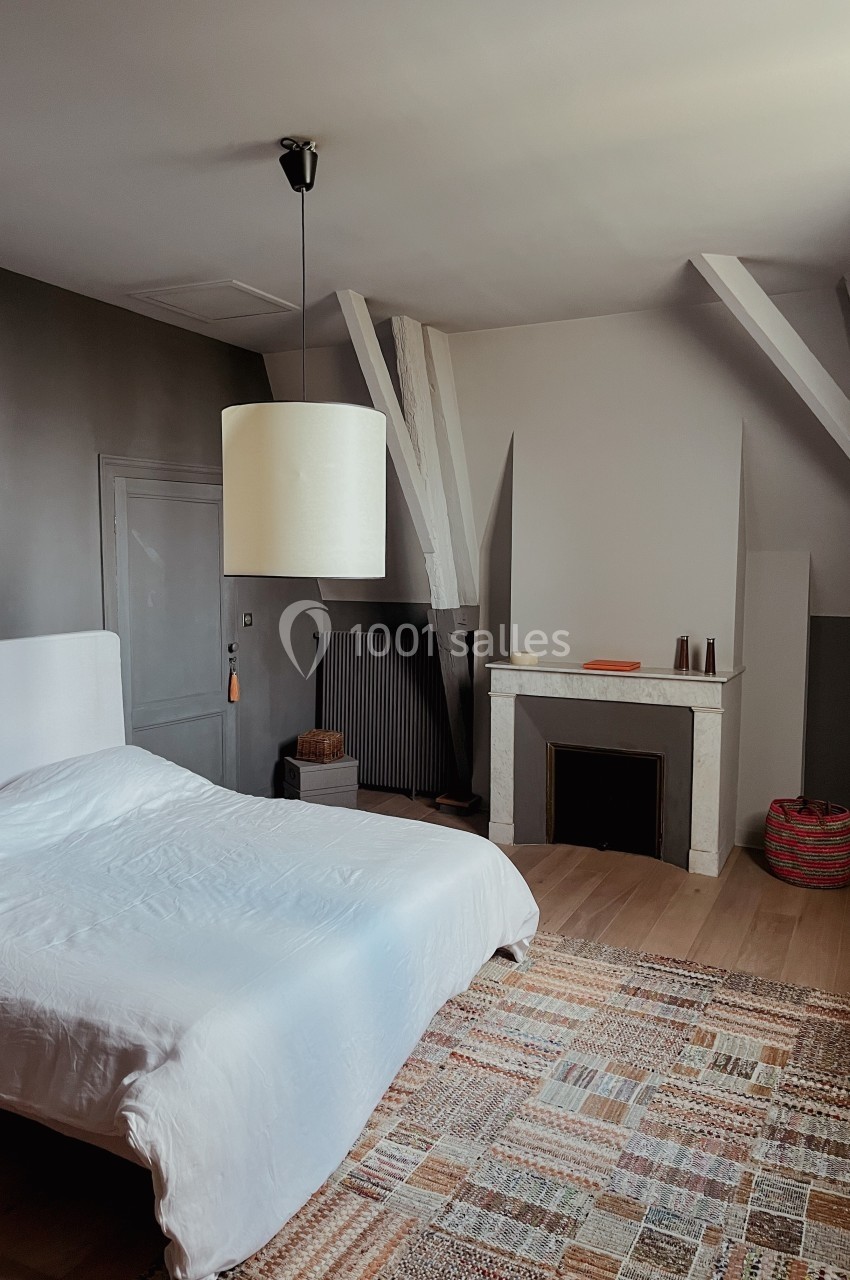 Chambre lumineuse avec lit double, cheminée décorative, tapis coloré et poutres apparentes sous un plafond mansardé.
