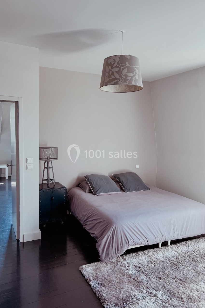 Chambre minimaliste avec lit double, murs beiges, parquet sombre, tapis clair et lampe suspendue.