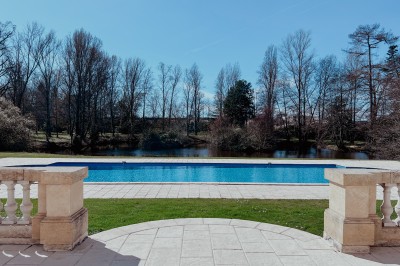 Vue sur une piscine extérieure entourée de pelouse, avec un lac et des arbres en arrière-plan sous un ciel dégagé.