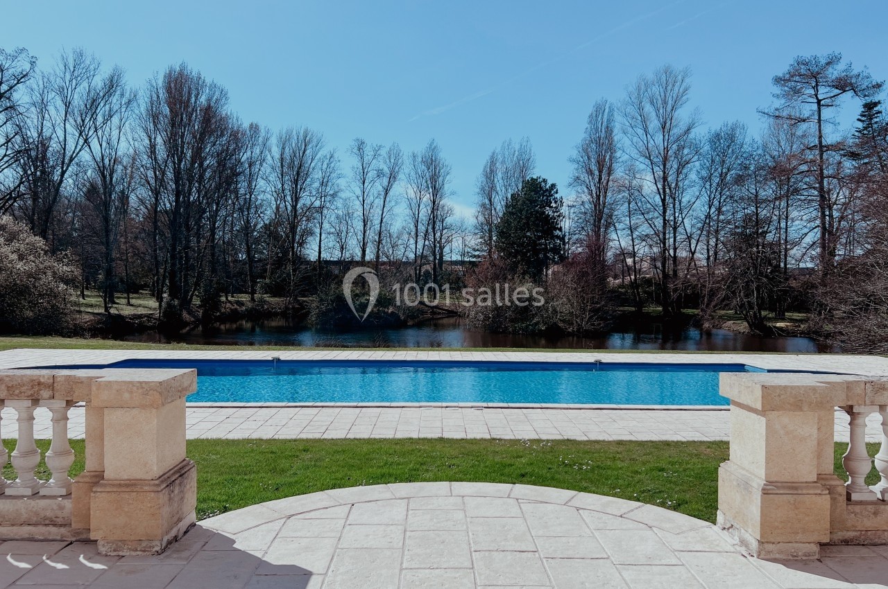 Vue sur une piscine extérieure entourée de pelouse, avec un lac et des arbres en arrière-plan sous un ciel dégagé.