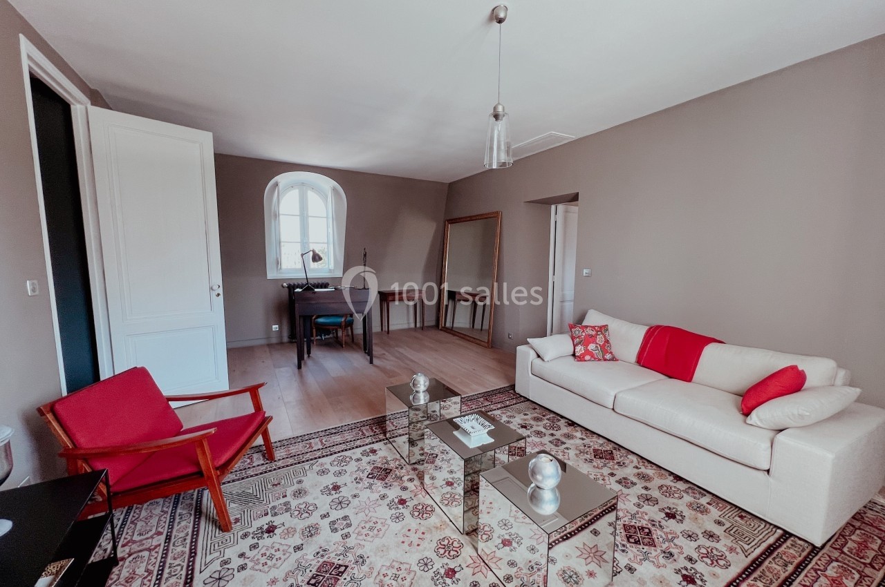 Salon lumineux avec canapé blanc, fauteuil rouge, tapis à motifs, bureau en bois et grande fenêtre cintrée.