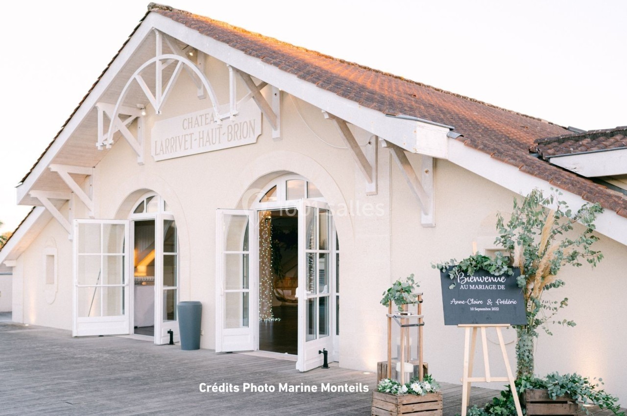 Façade d'un bâtiment clair avec des portes vitrées ouvertes, une terrasse en bois et un panneau décoratif à l'entrée.